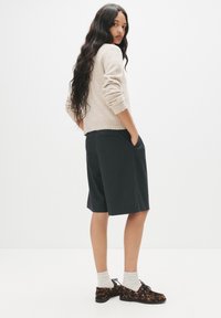 Maglione beige chiaro, pantaloni corti a righe scure e scarpe con motivo. I lunghi capelli cadono; le mani nelle tasche dei pantaloni.