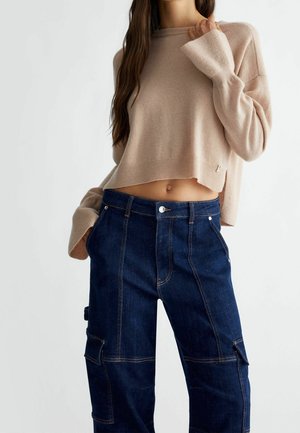 Woman wearing beige cropped sweater and dark blue high-waisted cargo jeans, with hands in pockets and one hand holding sweater collar.
