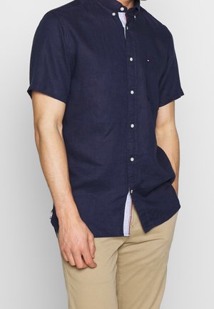Homme portant une chemise bleu marine à manches courtes boutonnée avec un petit logo sur la poitrine et un pantalon beige, la main reposant à ses côtés.