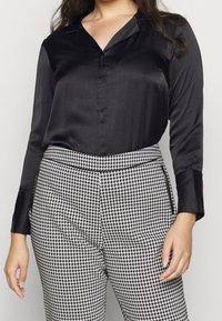 Blusa di raso nera con colletto a punta e maniche lunghe, abbinata a pantaloni a quadri houndstooth bianco e nero.