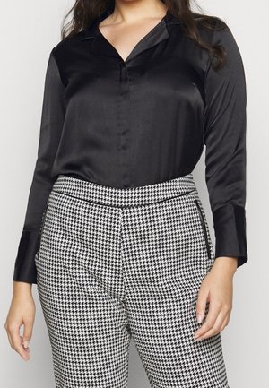 Schwarze Satinbluse mit spitzem Kragen und langen Ärmeln, kombiniert mit einer Hose im schwarz-weißen Hahnentritt-Muster.