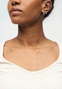 Približna snimka vrata i donjeg dijela lica osobe, prikazujući glatku kožu, neutralne usne, srebrne naušnice u obliku obruča i kremasti rub majice.