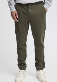 Olivgrüne Jogginghosen aus weichem Stoff mit einem elastischen Bund, versehen mit einem Kordelzug und seitlichen Taschen. Getragen mit weißen Turnschuhen.
