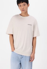 Beige bomull t-skjorte med korte ermer, rund halslinning, og liten svart "Club1990" tekst på brystet, sammen med lyseblå denimjeans.