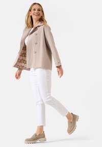 Helle beige Leinenjacke mit doppelter Knopfleiste und umgeschlagenen Ärmel, die das gemusterte Futter zeigt, kombiniert mit weißen Slim-Fit-Hosen und beigen Schuhen.