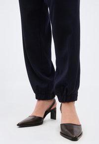 Γυναίκα που φοράει μαύρες μυτερές γόβες με λουράκι στο πίσω μέρος και χαλαρό σκούρο μπλε παντελόνι με ελαστικές μανσέτες, στέκεται πάνω σε λευκή επιφά νεια.