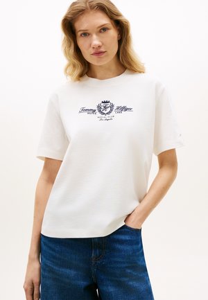 Ξανθιά γυναίκα με λευκό πουκάμισο Tommy Hilfiger με μπλε σκούρο λογότυπο και κείμενο, μπλε τζιν, στέκεται με το ένα χέρι στην τσέπη.