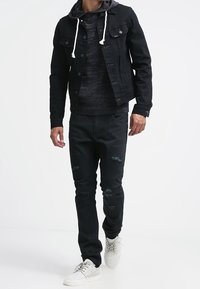 Černá džínová bunda s knoflíky a šedá mikina s kapucí pod ní, doplněné o potrhané černé džíny a bílé tenisky.