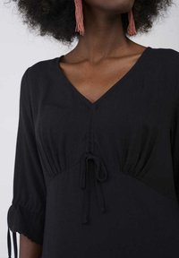 Svart klänning med V-ringning, rynkad midja och dragsko. Har trekvartslånga ärmar med vågiga kanter.