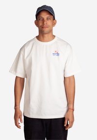 Hvid bomulds T-shirt med korte ærmer, rund halsudskæring og et lille logo med tekst og design i rød og blå på venstre bryst.