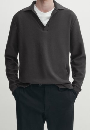 Personne portant un polo à manches longues gris foncé sur un t-shirt blanc avec un pantalon noir, les mains dans les poches, devant un fond uni.