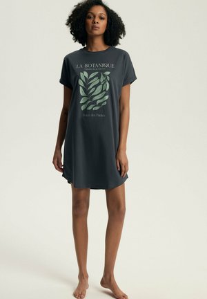 Kobieta o kręconych włosach, ubrana w ciemnoszarą sukienkę z krótkim rękawem z zielonym botanicznym nadrukiem i francuskim tekstem, stojąca boso na jednolitym tle.