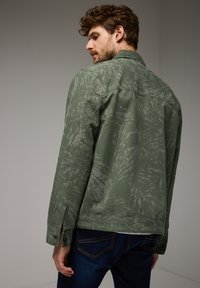 Zelená texturovaná bunda s jemným potiskem, s špičatým límcem a knoflíkovými manžetami, v kombinaci s tmavě modrými džíny.