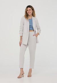 Blazer grigio chiaro su misura con un solo bottone, abbinato a pantaloni grigi attillati. Sotto, un top corto blu. Tacchi beige con cinturini.