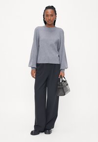 Hallide hall sweater, millel on laiad varrukad, koos mustade laia säärega pükstega. Mudel hoiab käes väikest musta käekotti, millel on tekstuuriga muster.