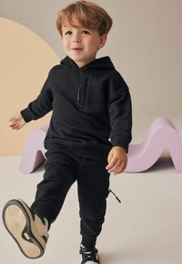 Sweatshirt à capuche noir avec poche avant, associé à un jogger assorti avec une poche zippée. L'ensemble est complété par des baskets noires.
