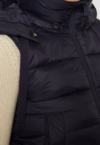 Marinblå puffervest med hög krage, dragkedjestängning och tryckknappar. Har en framficka och justerbara remmar på axlarna.