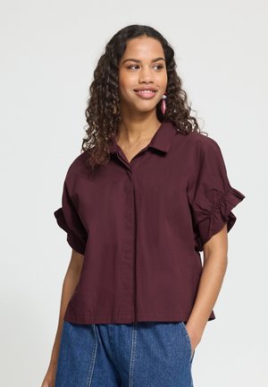 Junge Frau mit lockigem Haar, die eine kurzärmelige, dunkelviolette Bluse und blaue Jeans trägt, lächelnd und zur Seite schauend.