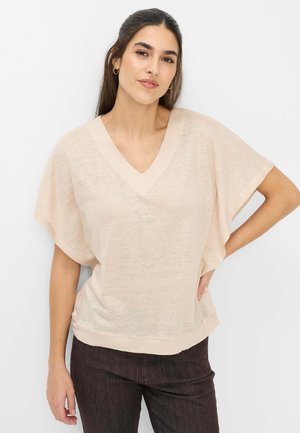 Junge Frau mit langen dunklen Haaren trägt ein beiges V-Ausschnitt Kurzarmshirt und dunkle Hosen, posiert mit einer Hand in der Hüfte vor weißem Hintergrund.