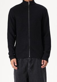 Svart ribbad stickad zip-up jacka med hög krage, som har långa ärmar och en figursydd midja, tillverkad av texturerat tyg för extra värme.