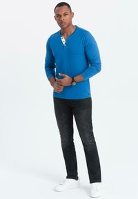 Blauwe langarmsgemerkte henley shirt met een knoopsluiting bij de hals, gecombineerd met zwarte jeans en witte sneakers. Het shirt heeft een gladde textuur.