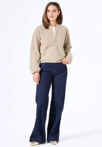Een tan fleece bomberjack met een ronde hals en een rits aan de voorkant, gecombineerd met donkerblauwe flared jeans en lichtgekleurde schoenen.