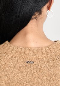 Personne avec de longs cheveux noirs portant un pull en tricot beige affichant une étiquette argentée "BOSS" dans le haut du dos et une boucle d'oreille en argent.