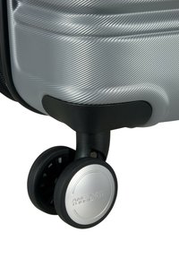 Angolo di una valigia rigida argentata con finitura testurizzata, caratterizzata da una base nera per le ruote e da un mozzo per le ruote argentato metallizzato con branding.