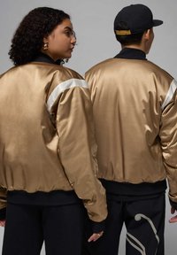 Giubbotti bomber in satin dorato con accenti a strisce bianche, polsini a coste e colletto nero, visti da dietro. Sotto sono indossati pantaloni neri.