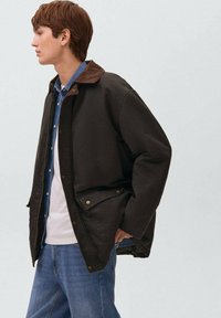 Giacca in cotone marrone con colletto in velluto a coste, chiusure a scatto e tasche frontali, indossata su una camicia blu e una t-shirt bianca, con jeans blu.