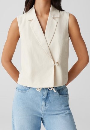 Mouwloze crèmekleurige blazer met enkele knoopsluiting, gedragen over lichtblauwe, hooggetaillede jeans met een koordsluiting in de taille.