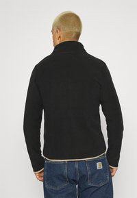 Czarna polarowa kurtka z wysokim kołnierzem, teksturowana tkanina i kremowe wykończenie u dołu oraz na mankietach. Połączona z niebieskimi dżinsami.