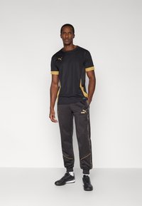 Musta urheilupaita kultaisilla yksityiskohdilla, yhteensopivien harmaiden vetohousujen kanssa, joissa on kullanväriset raidat ja logo. Malli seisoo urheilukengissä.