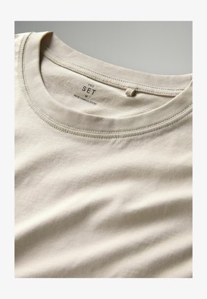 Beige Baumwoll-T-Shirt mit rundem Rundhalsausschnitt und weicher Textur. Das Etikett trägt die Aufschrift "THE SET M" und weist darauf hin, dass es "MADE IN BANGLADESH" ist.
