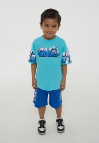 T-shirt azzurro a maniche corte con testo grafico; pantaloni corti blu con design simile. Indossato con sneakers nere e dettagli bianchi.