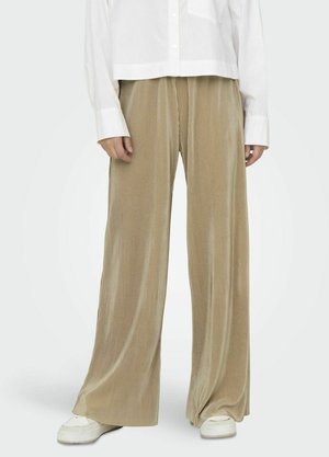 Beige, weit geschnittene, gerippte Hosen aus einem weichen Material. Kombiniert mit einer weißen, kurz geschnittenen Bluse mit Knöpfen und weißen Sneakern.