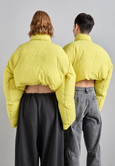 Yellow padded jackets with a cropped design feature a textured finish and embellishments. Worn over dark gray pants, seen from the back.