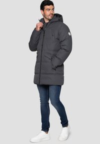 Mörkgrå pufferjacka med huva, med en framgående dragkedja, knäppt bröstficka och sidofickor. I kombination med blå jeans och vita sneakers.