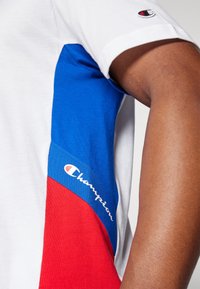 Camiseta blanca de manga corta con paneles de tela azules y rojos. Presenta el logo "Champion" bordado en blanco en el panel azul. Textura suave.