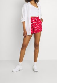 Shorts rossi a fiori con cintura annodata, abbinati a un crop top bianco e una camicia bianca aperta. Indossati con sneakers bianche e calze.