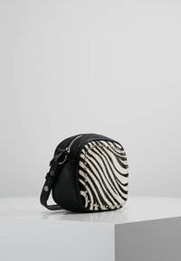 Kulatá kabelka přes rameno z černé umělé kůže a zebrované látky, zdobená stříbrnými nýty podél okraje a se zapínáním na zip.
