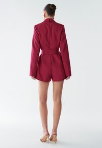 Burgundský prispôsobený overal s dlhými rukávmi, opaskom v páse a krátkymi spodkami, vyrobený z hladkej látky. Viditeľný zips na chrbte.