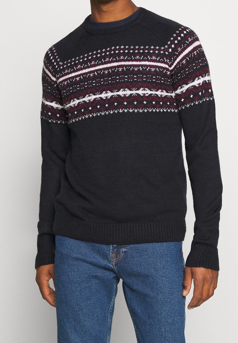 Homme portant un pull bleu marine avec des bandes rouges, blanches et roses à motifs sur la poitrine et les parties supérieures des manches, associé à un jean bleu.