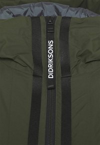 Oliivinvihreä takki, jossa on musta vetoketju ja hihnat. Varustettu korkealla kauluksella ja ommelluilla saumoilla. "DIDRIKSONS" -logo hihnassa. Kevyt materiaali.