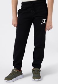 Zwarte fleece joggerbroeken met elastische tailleband, voorzien van een wit Champion-logo en zijzakken, gecombineerd met groene sportschoenen.