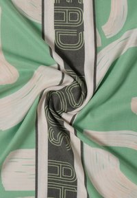 Tessuto verde con motivi astratti di pennellate chiare, sovrapposte a forme geometriche e scritte nere audaci. Texture morbida.