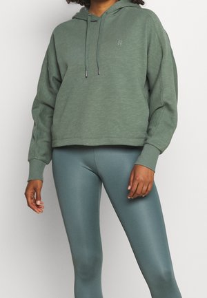 Sudadera verde con un corte relajado y capucha con cordones, combinada con leggings de color teal suave. Ambas prendas cuentan con una textura suave.