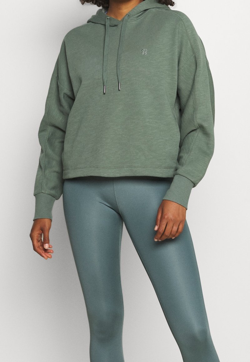 Felpa verde con una vestibilità comoda e cappuccio con cordino, abbinata a leggings teal lisci. Entrambi i capi presentano una texture morbida.