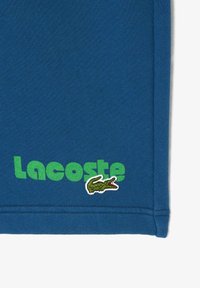 Tela de algodón azul con la palabra "Lacoste" en verde y un logotipo de cocodrilo verde. El diseño tiene una textura suave con bordes cosidos.