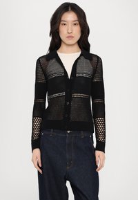 Black knit cardigan with a structured design, featuring a textured mesh pattern, button closure, and collar. Worn over a light top and dark jeans.
